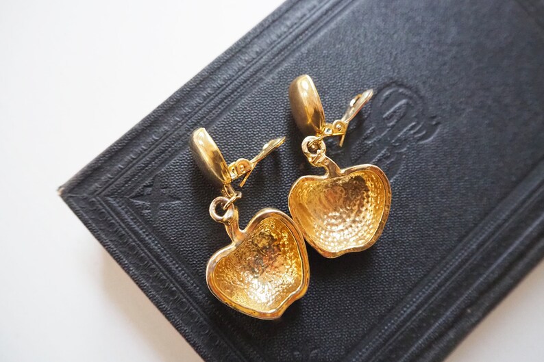 May include: A pair of gold-tone clip-on earrings featuring a detailed apple design. The earrings are made of metal and have a shiny finish.