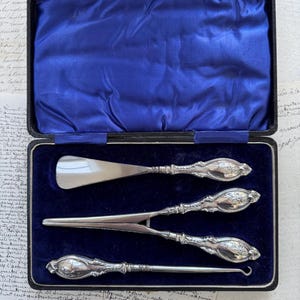 Peut inclure: Un ensemble de trois outils en argent pour chausse-pieds dans une boîte doublée de velours bleu. Les outils comprennent un chausse-pied, un crochet à boutons et un crochet à lacets.