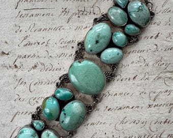 Op de afbeelding: Een zilverkleurige armband met meerdere ovale en afgeronde turkooizen stenen. De stenen zijn gezet in een decoratieve, geschakelde metalen band. De armband wordt getoond op een vintage papieren achtergrond met handschrift.