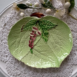 Puede incluir: Un plato de cerámica verde con forma de hoja con un diseño de flores rosas y blancas. El plato está sobre un encaje blanco.