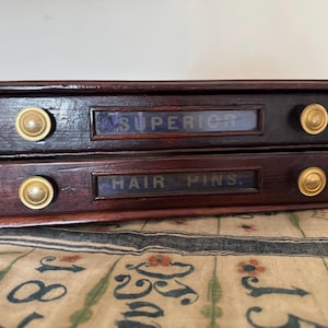 Puede incluir: Una caja de almacenamiento de madera oscura de dos cajones con pomos de latón. El cajón superior dice "SUPERIOR" y el cajón inferior "HAIR PINS". La caja está sobre una tela estampada.