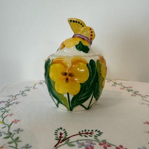 Może przedstawiać: Biały ceramiczny dzbanek z żółtym motylem na pokrywce. Dzbanek jest ozdobiony żółtymi i zielonymi fiołkami.