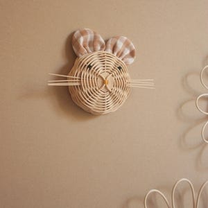 May include: A woven rattan bunny head wall decoration with plaid fabric ears and whiskers. The bunny's eyes are small black beads, and the nose is a small yellow bead. The wall decor is a light beige color and is mounted on a beige wall.