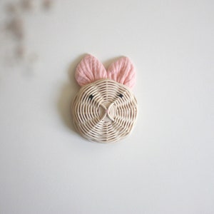 May include: A woven wicker bunny head wall decor with pink fabric ears and black eyes. The bunny is round and has a simple, minimalist design, perfect for a nursery or children's room. The bunny is mounted on a white wall.
