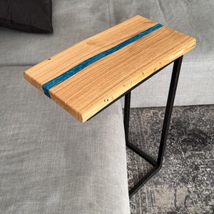 Könnte beinhalten: Ein Beistelltisch aus Holz mit rechteckiger Platte und blauem Harzeinlass. Der Tisch hat ein schwarzes Metallgestell und ist so konzipiert, dass er über die Armlehne eines Sofas geschoben werden kann. Das Holz hat eine natürliche Maserung.