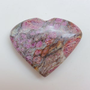 Può includere: Una pietra a forma di cuore con una miscela di tonalità rosa, grigie e marroni. La superficie presenta un aspetto strutturato con venature e motivi visibili. L'oggetto è probabilmente un elemento decorativo o un gioiello.