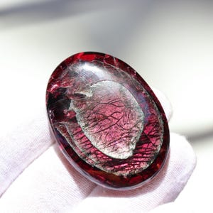 May include: A polished, oval-shaped gemstone with a deep red hue, featuring internal layers and a shimmering, silvery-gray pattern. The stone is held against a white background, highlighting its translucent quality and intricate details.