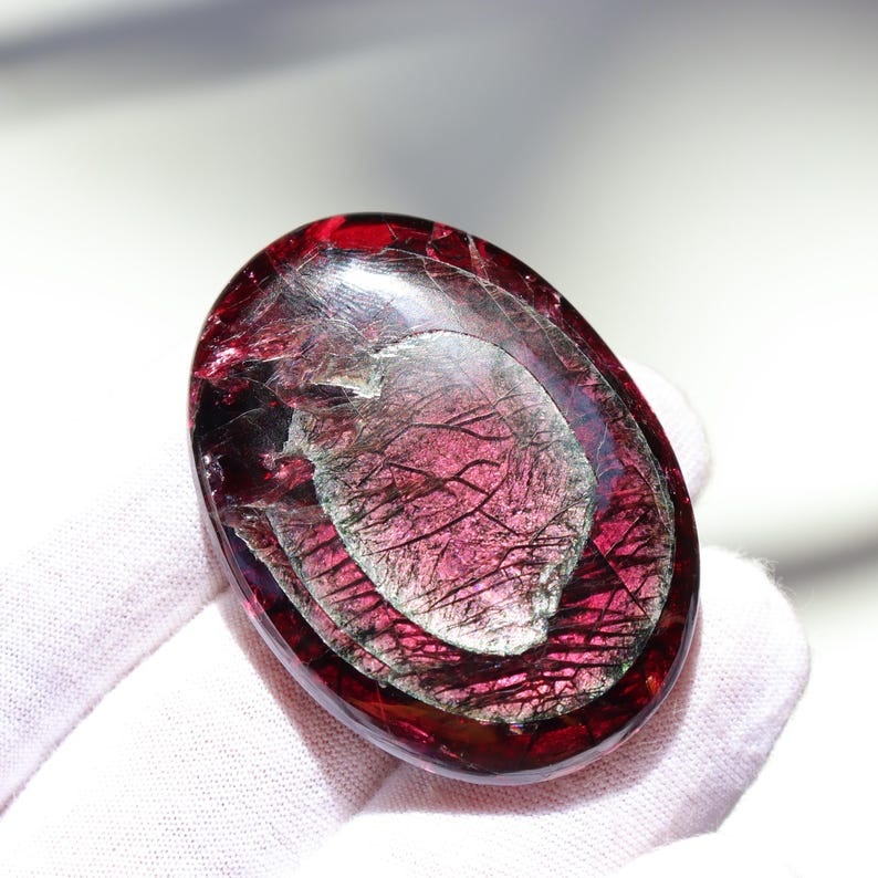 May include: A polished, oval-shaped gemstone with a deep red hue, featuring internal layers and a shimmering, silvery-gray pattern. The stone is held against a white background, highlighting its translucent quality and intricate details.