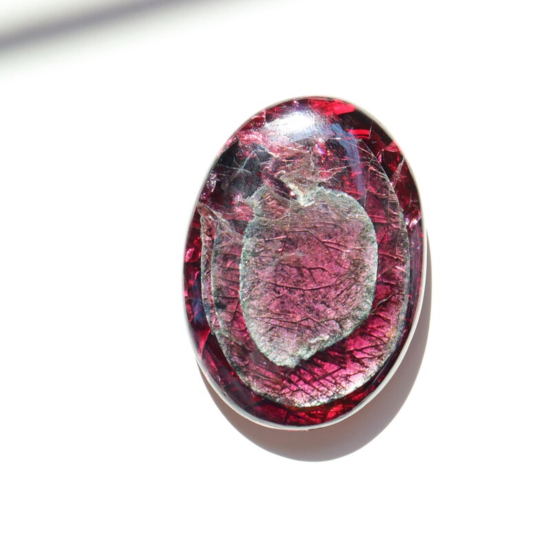 May include: An oval-shaped, dichroic glass pendant with a silver-colored metal border. The pendant features layers of deep red and purple glass, with a central, leaf-like design in shades of silver and gray. The glass has a textured, crackled appearance.