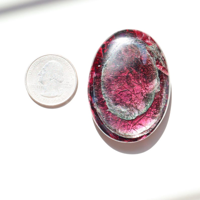 May include: An oval-shaped, polished gemstone with a deep magenta hue, featuring internal fractures and a layered appearance. A U.S. quarter coin is next to the gemstone for size comparison. The gemstone has a translucent quality.