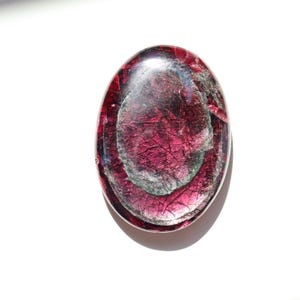 May include: An oval-shaped, polished gemstone with a deep magenta hue, featuring internal patterns of silver and darker magenta. The stone has a glossy, reflective surface, and is set against a white background.