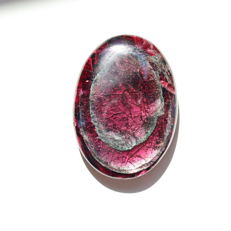 May include: An oval-shaped, polished gemstone with a deep magenta hue, featuring internal patterns of silver and darker magenta. The stone has a glossy, reflective surface, and is set against a white background.