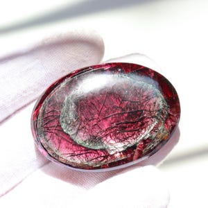 May include: An oval-shaped, polished gemstone with a deep red hue and internal fractures. The stone has a translucent quality, revealing layers of color and texture. The gemstone is held in a white glove.