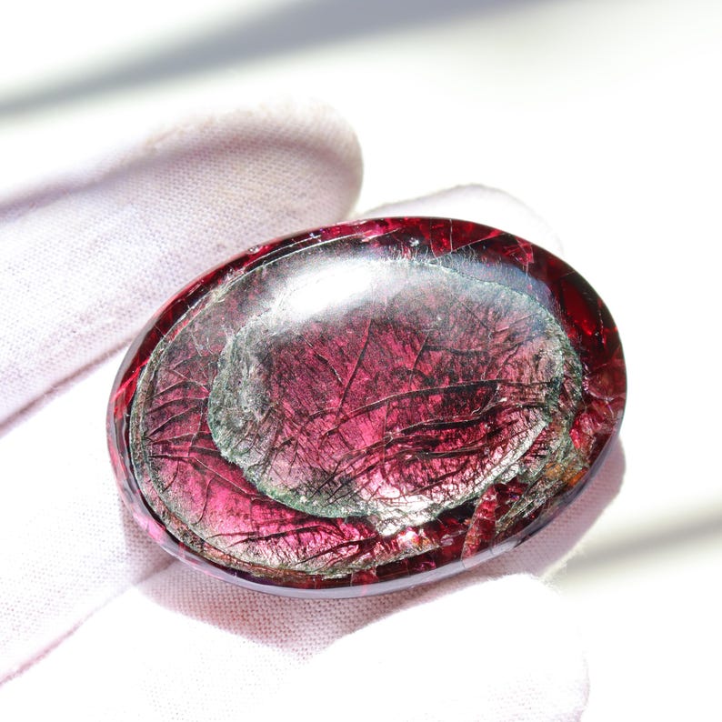 May include: An oval-shaped, polished gemstone with a deep red hue and internal fractures. The stone has a translucent quality, revealing layers of color and texture. The gemstone is held in a white glove.