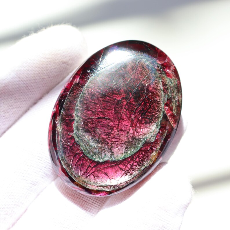 May include: An oval-shaped, polished gemstone with a deep burgundy hue, featuring internal patterns of lighter red and green. The stone has a glossy surface with visible internal fractures, creating a unique, textured appearance. The gemstone is held by a gloved hand.