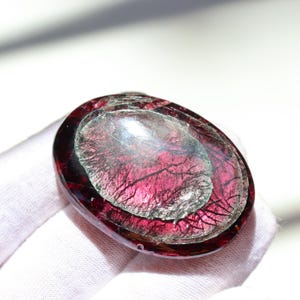 May include: Oval-shaped, polished gemstone with a deep red outer layer and a lighter, translucent center. The gemstone has intricate, vein-like patterns throughout, creating a unique visual texture. The stone is held in a white gloved hand.