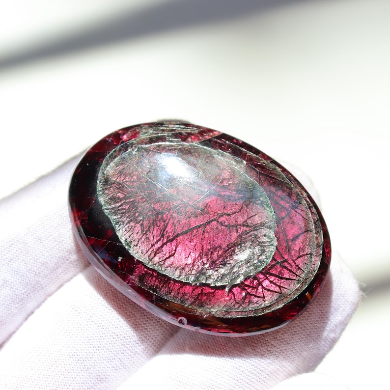 May include: Oval-shaped, polished gemstone with a deep red outer layer and a lighter, translucent center. The gemstone has intricate, vein-like patterns throughout, creating a unique visual texture. The stone is held in a white gloved hand.