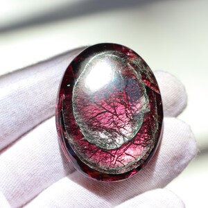 May include: Oval-shaped gemstone with a deep red hue, featuring internal layers and intricate veining. The polished surface reflects light, creating a shimmering effect. The gemstone is held by a gloved hand.
