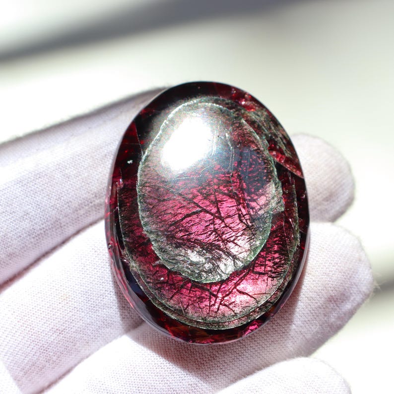 May include: Oval-shaped gemstone with a deep red hue, featuring internal layers and intricate veining. The polished surface reflects light, creating a shimmering effect. The gemstone is held by a gloved hand.