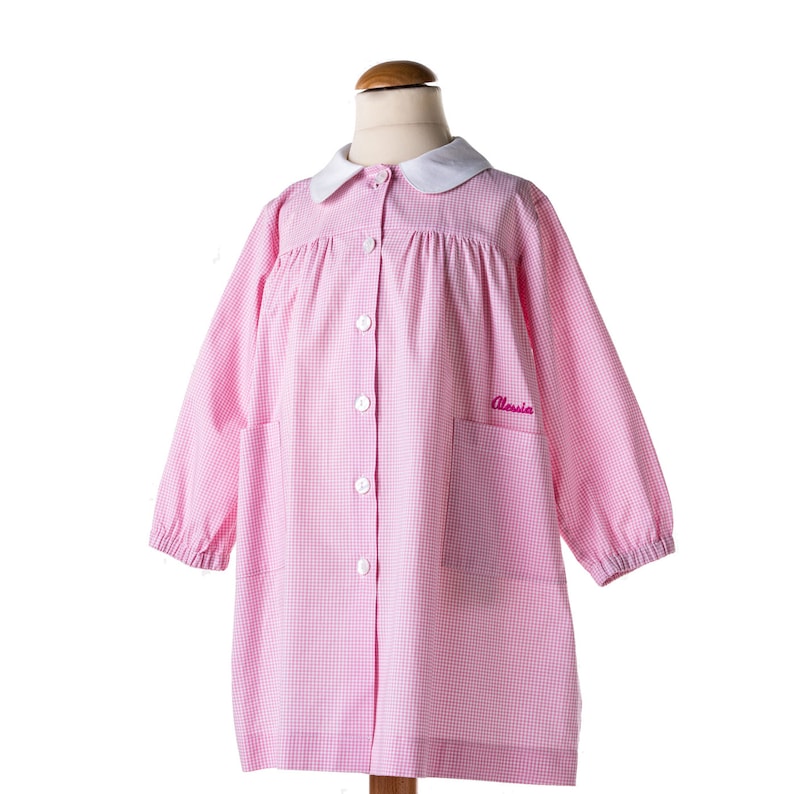 Puede incluir: Smock de vichy rosa y blanco con cuello blanco y mangas largas. La smock tiene cierre de botones y un bolsillo en el lado izquierdo. El nombre "Alessia" est&aacute; bordado en el bolsillo en rosa.