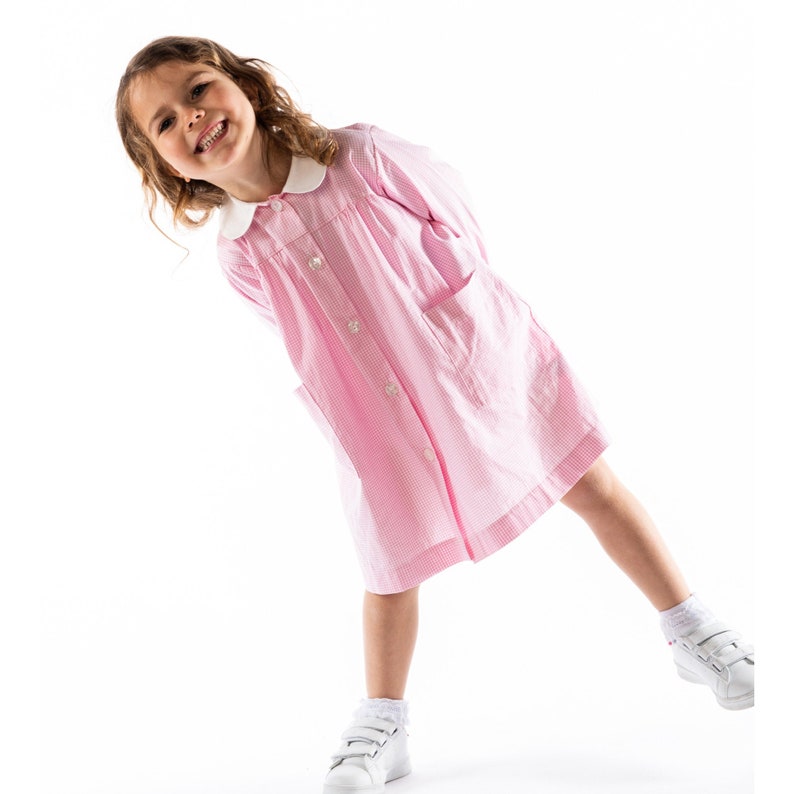 Puede incluir: Una ni&ntilde;a peque&ntilde;a con un vestido de cuadros rosa y blanco con cuello blanco y botones. Est&aacute; sonriendo y tiene las manos en los bolsillos.