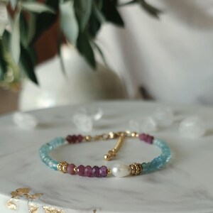 Peut inclure: Un bracelet délicat composé de perles facettées bleu clair aigue-marine, de perles rose foncé aux tons rubis et d'une perle d'eau douce blanche centrale. Le design inclut de petites entretoises en métal doré et un fermoir à chaînette.