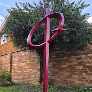 Puede incluir: Escultura de metal roja para exteriores con diseño de estrella y óvalo. La escultura se encuentra en un jardín con césped frente a una pared de ladrillos y un arbusto verde. El cielo está nublado.