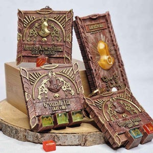 May include: Four intricately designed, rectangular, brown and gold decorative boxes. Each box features embossed details, including text and ornate embellishments. The boxes have a weathered appearance and are arranged on a wooden surface. Some boxes have small, colorful, square-shaped objects.