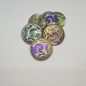 May include: A selection of decorative round buttons in shades of green, purple, and pink. Each button has an ornate, swirling design in gold or silver, with a metallic finish. The buttons are arranged on a white surface.