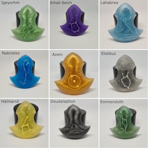 Op de afbeelding: Negen hars hangers in verschillende kleuren, elk met een uniek sterrenbeeld ontwerp en naam. Kleuren zijn groen, paars, blauw, goud, grijs en geel. De hangers worden op zwarte standaards gepresenteerd.