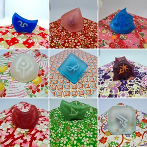 May include: Nine colorful, translucent resin objects with various symbols, including a crescent moon, sun, and other geometric shapes. Each object is a different color, such as blue, red, and green. They are displayed on patterned paper.