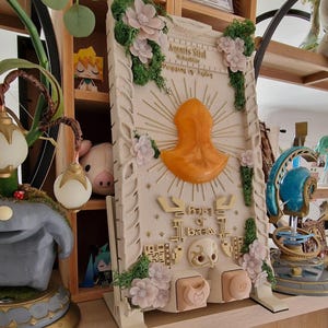 May include: A detailed, off-white decorative panel with an orange central design and gold accents. The panel features floral embellishments, green moss, and text. The panel is supported by a stand and is likely a decorative item.