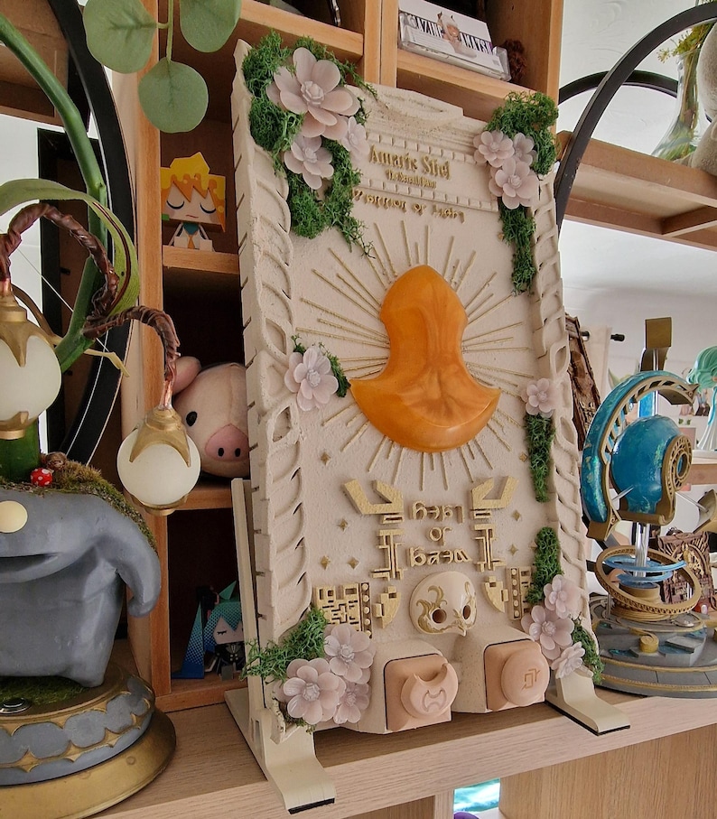 May include: A detailed, off-white decorative panel with an orange central design and gold accents. The panel features floral embellishments, green moss, and text. The panel is supported by a stand and is likely a decorative item.