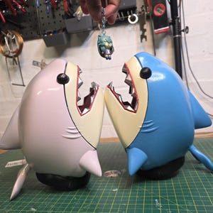 May include: Two shark-shaped sculptures, one light pink and the other blue, with open mouths revealing red and silver teeth. A small figurine hangs above them. The sculptures have black eyes and are set on a green cutting mat.