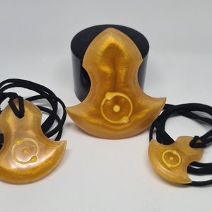 Op de afbeelding: Drie goudkleurige hangerkettingen met zwarte koorden. Elke hanger heeft een unieke vorm met een cirkelvormig ontwerp in het midden. De hangers worden gepresenteerd tegen een witte achtergrond.