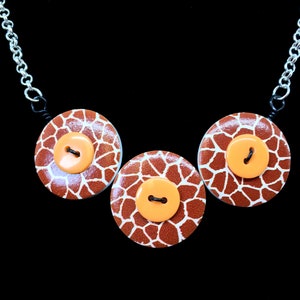 May include: A necklace with three circular pendants, each displaying a giraffe print pattern and an orange button. The chain is silver-coloured. The background is black, creating a striking contrast.