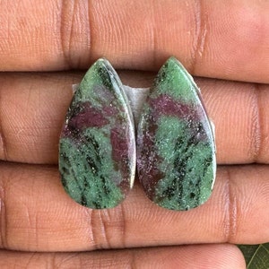 Może przedstawiać: Dwa kamienie szlachetne w kształcie łezki. Każdy kamień ma mieszankę kolorów zielonego i bordowego, z wypolerowaną powierzchnią. Kamienie szlachetne są osadzone na białym podłożu i trzymane w dłoni.