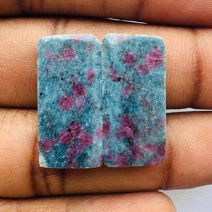 以下が含まれることがあります： 青とピンクのルビーの宝石の四角い形をした2つのピース。石は滑らかで平らな表面にカットされ、研磨されています。石はピンクと白のインクルージョンで斑点があります。