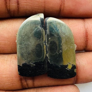 May include: Two polished, arched gemstones with a metallic sheen, held in a hand. The stones have a dark base and a lighter, reflective top. The gemstones are likely for jewelry or decorative purposes.