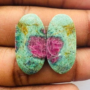 May include: Two oval-shaped green gemstones with pink and gold inclusions. The stones are set in a pair of earrings.