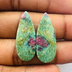 May include: Two teardrop-shaped green gemstones with pink and gold inclusions. The stones are set in a pear shape and have a smooth, polished finish.