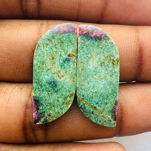 Könnte beinhalten: Zwei grüne und rosafarbene Edelsteinkabochons, jeweils mit einer abgerundeten Halbmondform. Die Steine haben ein geflecktes, gesprenkeltes Aussehen mit grünen, rosafarbenen und braunen Farbtönen.