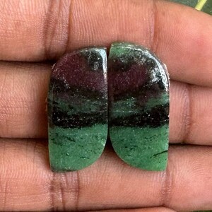 Puede incluir: Dos piedras preciosas de zoisita pulidas, cada una con un patrón único de bandas rojas oscuras, negras y verdes. Las piedras son lisas y redondeadas, ideales para la fabricación de joyas o coleccionar. Las piedras preciosas se sostienen en una mano.