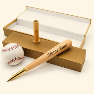 Puede incluir: Un bolígrafo de madera clara con forma de bate de béisbol en miniatura con el texto Home Run! grabado en el lateral, con una punta de metal dorado y una tapa de madera a juego, presentado junto a una pelota de béisbol blanca con costuras rojas.