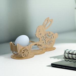 May include: Wooden Easter bunny figurine holding a white egg in a small wagon. The bunny is intricately carved with floral details.