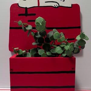 May include: A red wooden planter shaped like Snoopy's doghouse, with black stripes and a white Snoopy figure on top. The planter is filled with green leafy plants. A charming, decorative item.