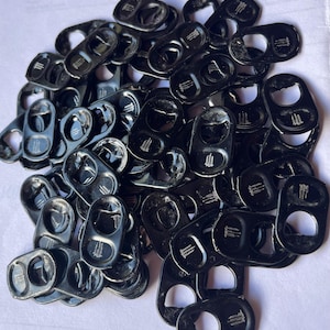 May include: A pile of black metal can pull tabs with a glossy finish. Each tab has a circular opening and a rectangular section with a logo. The tabs are scattered on a light-colored surface.