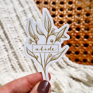 May include: A white sticker with a gold outline of a floral bouquet. The word "abide" is written in gold script inside a rectangular box.