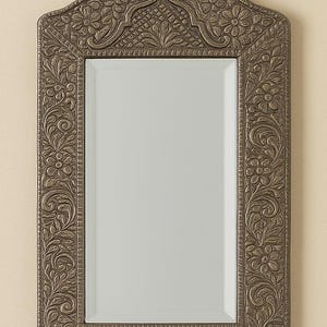 May include: Ornate rectangular mirror with a decorative, floral-patterned frame in a bronze finish. The mirror has a slightly arched top and is designed for wall hanging. The frame features detailed carvings of flowers and leaves, adding a touch of elegance.