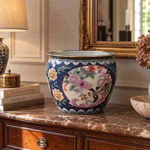 Pode incluir: Um vaso de cerâmica redondo com um fundo azul profundo e padrões florais dourados e brancos intrincados. O centro exibe um grande medalhão em forma de leque de cor creme decorado com peónias cor-de-rosa, folhagem verde e pequenos pássaros.